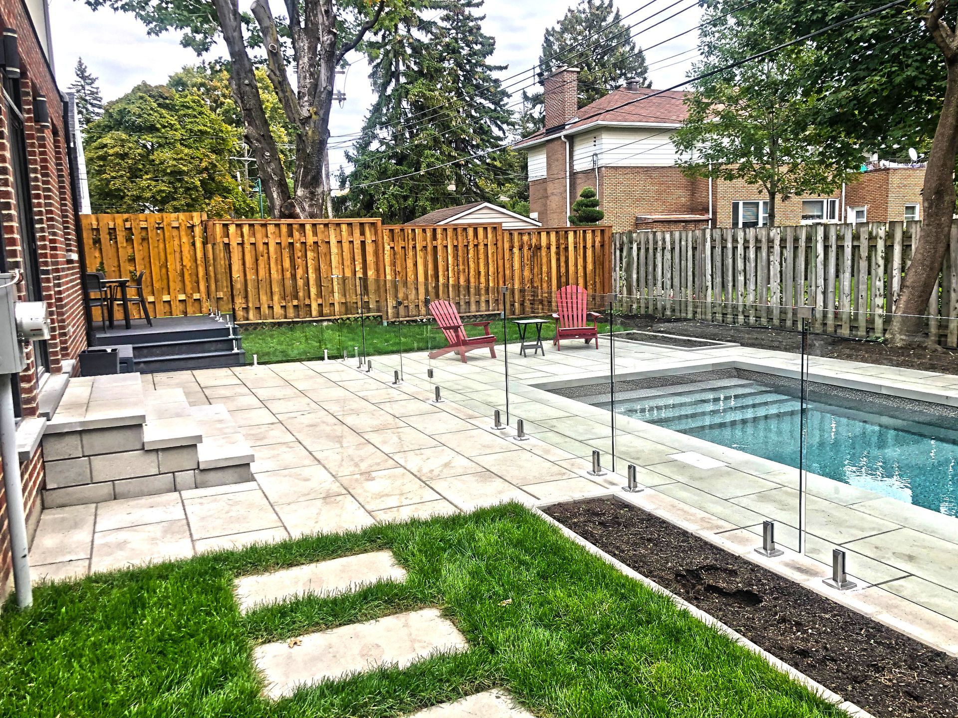 Terrasse arrière avec piscine, pelouse et clôture en bois. Deux chaises rouges et un jacuzzi sont installés sur une surface pavée.