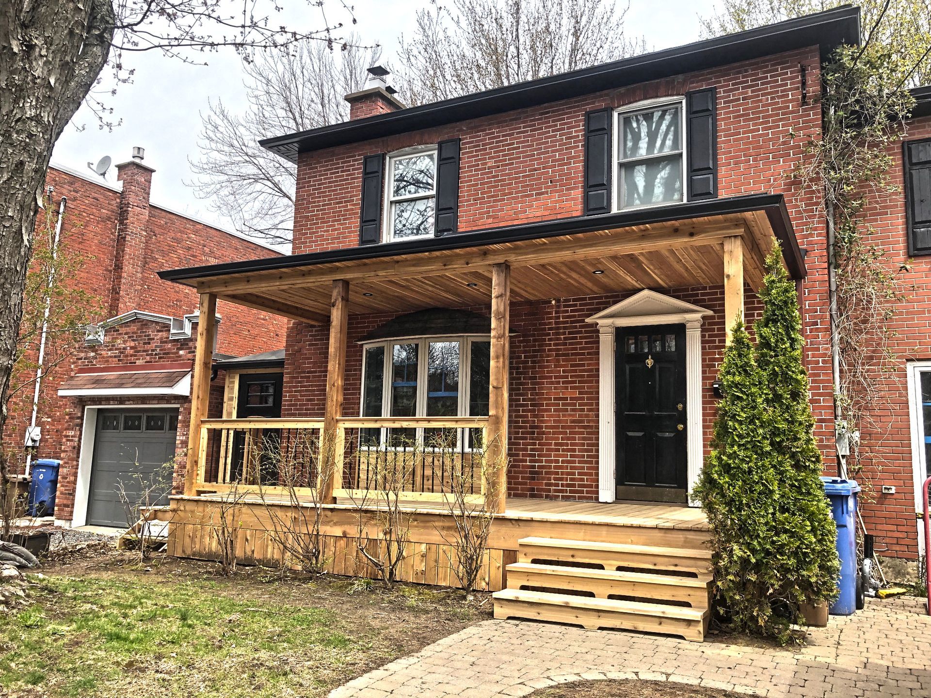 Red brick house with porch, black shutters, and black door. Green shrubbery to the right.