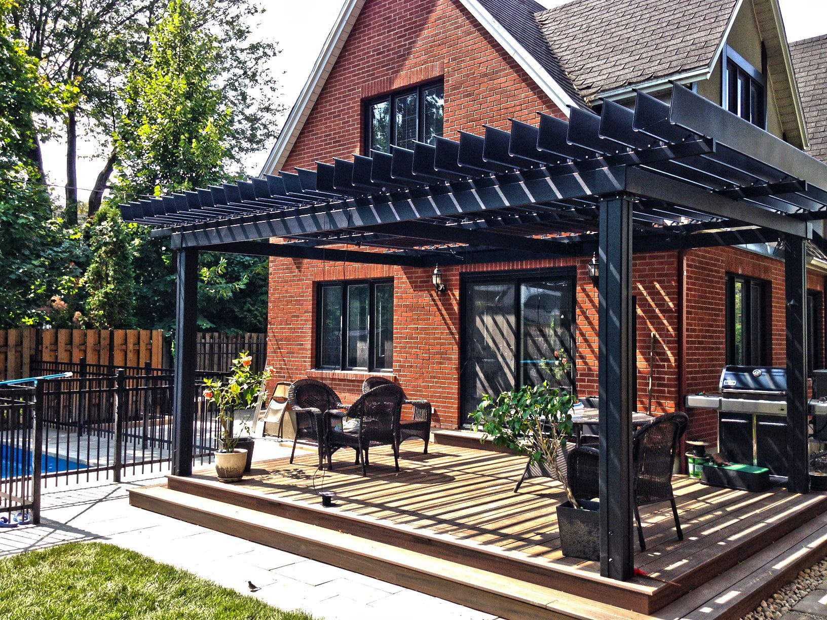 Black pergola over a wooden deck with seating, adjacent to a brick house and swimming pool.