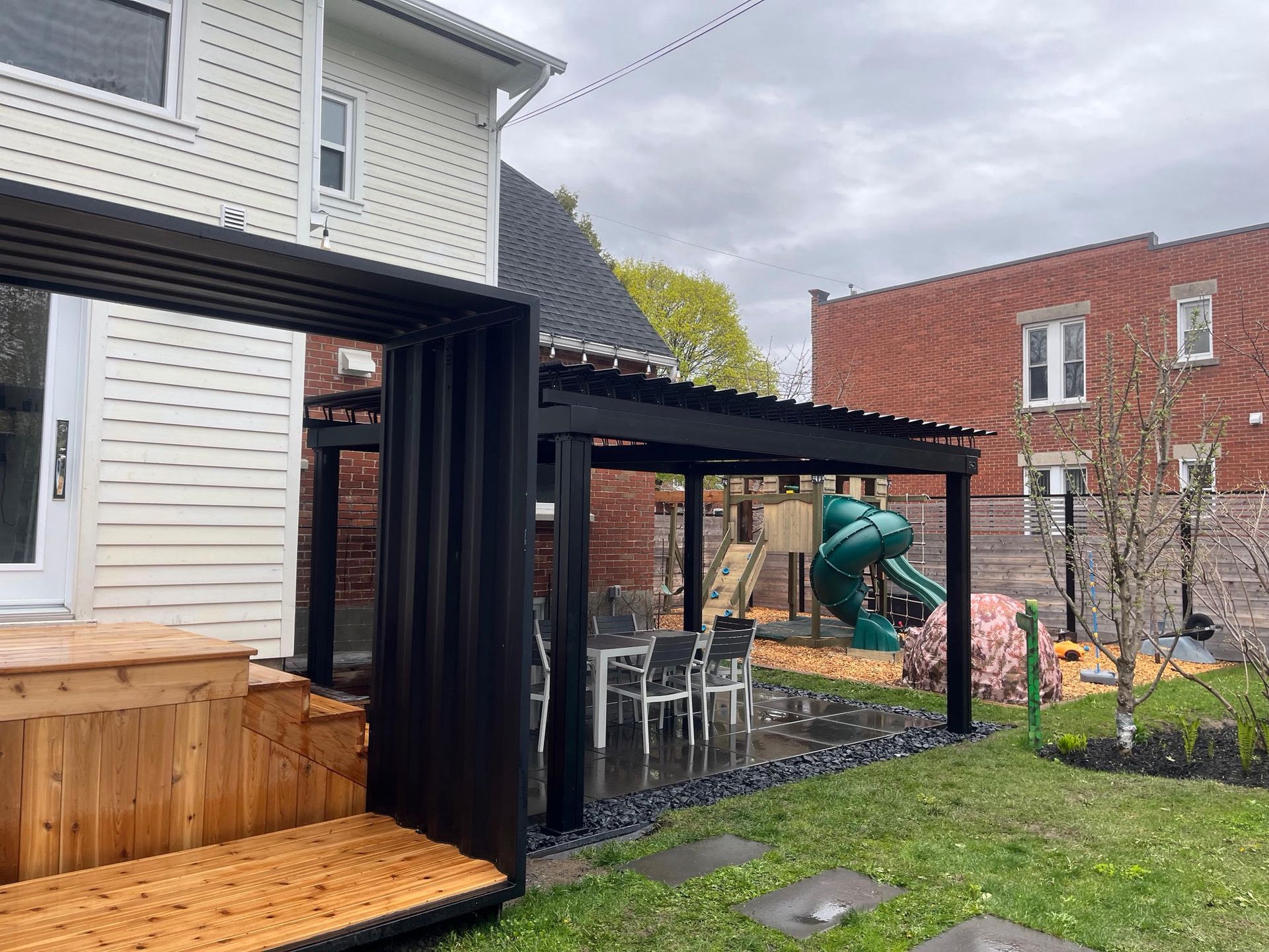 Black pergola with dining set on a lawn next to a wood deck, a house and a play structure.