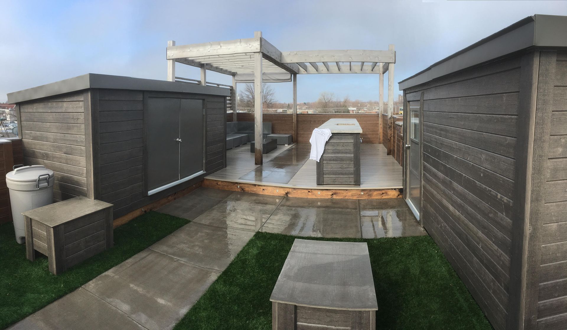 Rooftop deck with gray structures, pergola, and artificial grass. A wet bar, storage sheds, and a pathway are visible.