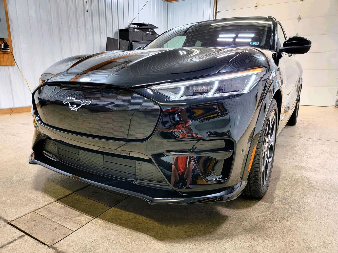Black Ford Mustang Mach-E electric SUV. Front view, showing headlights, grille, and sleek design in a garage setting.