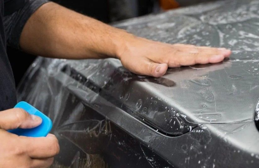 Person smoothing a protective film on a car's hood with a blue applicator.