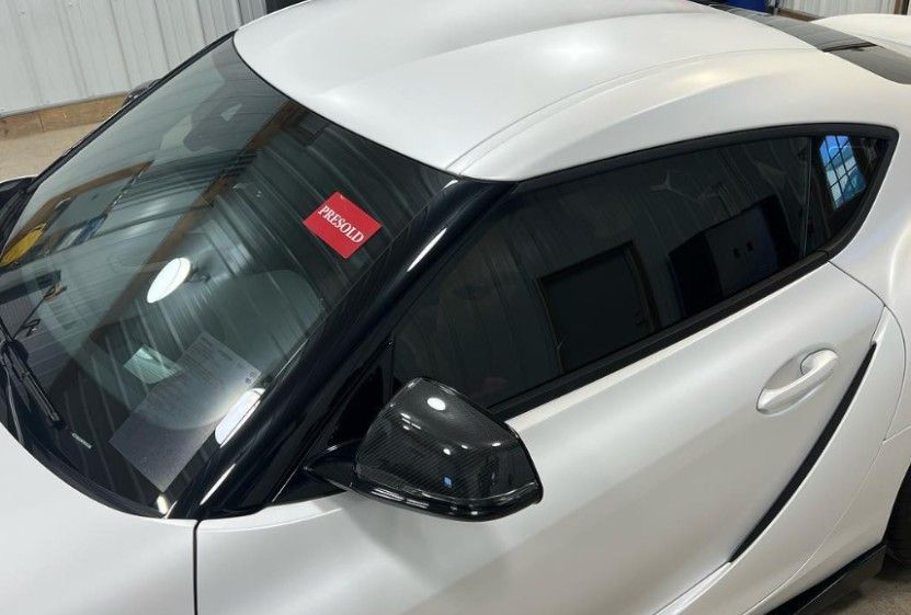 White sports car with tinted windows, black trim, and carbon fiber side mirror. Sold sticker visible.