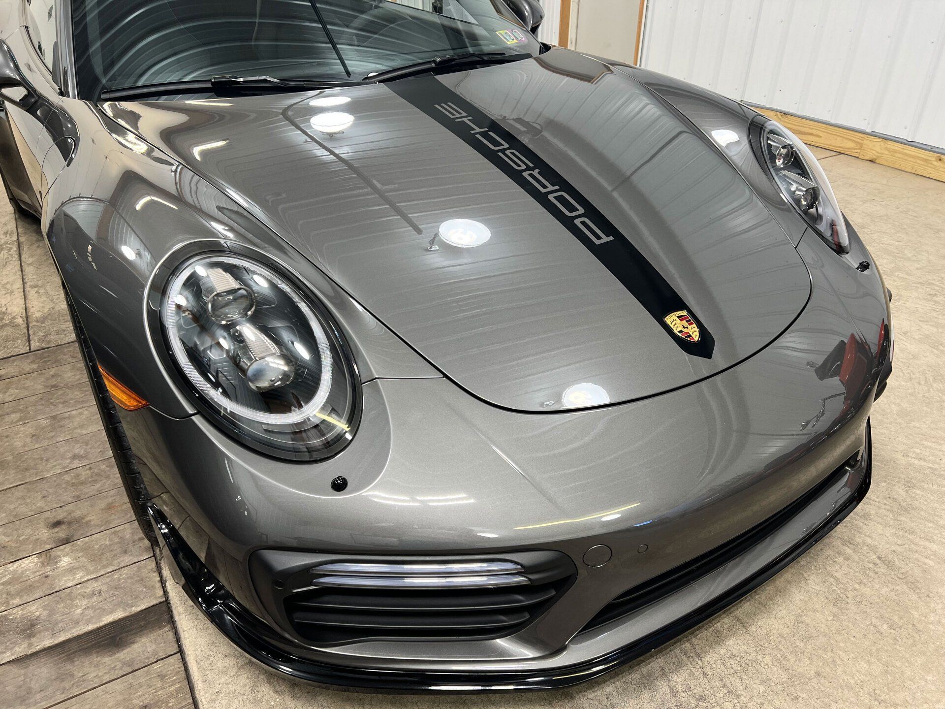 Gray Porsche 911 front view in a garage, showing the hood, headlights, and black racing stripe