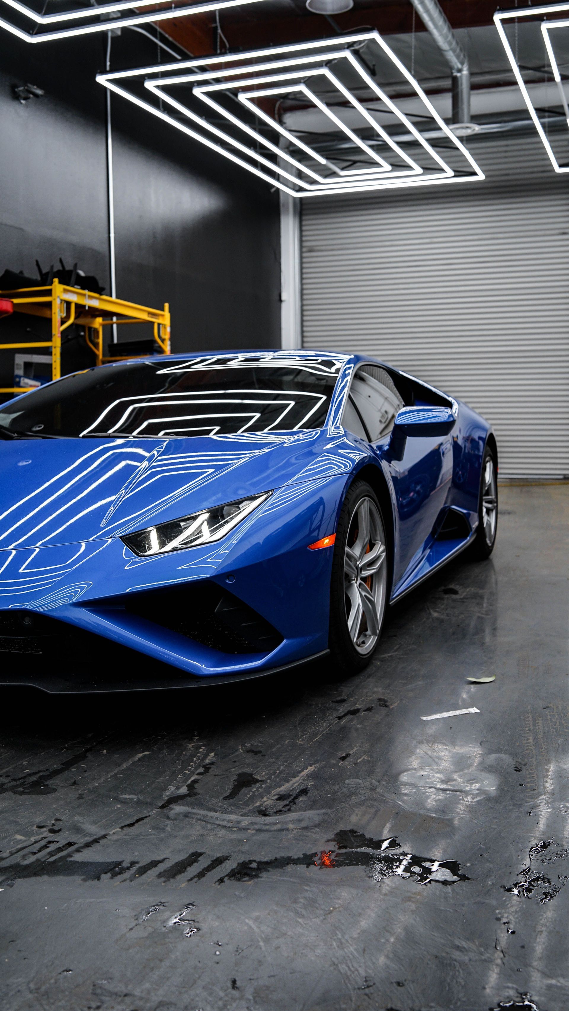Blue sports car in a sleek garage under geometric ceiling lights, shown front-left on a wet floor.