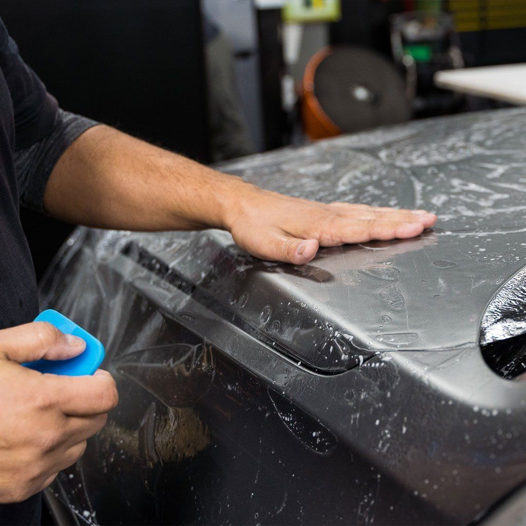 Hands applying a clear protective film to a dark car hood with a blue tool.
