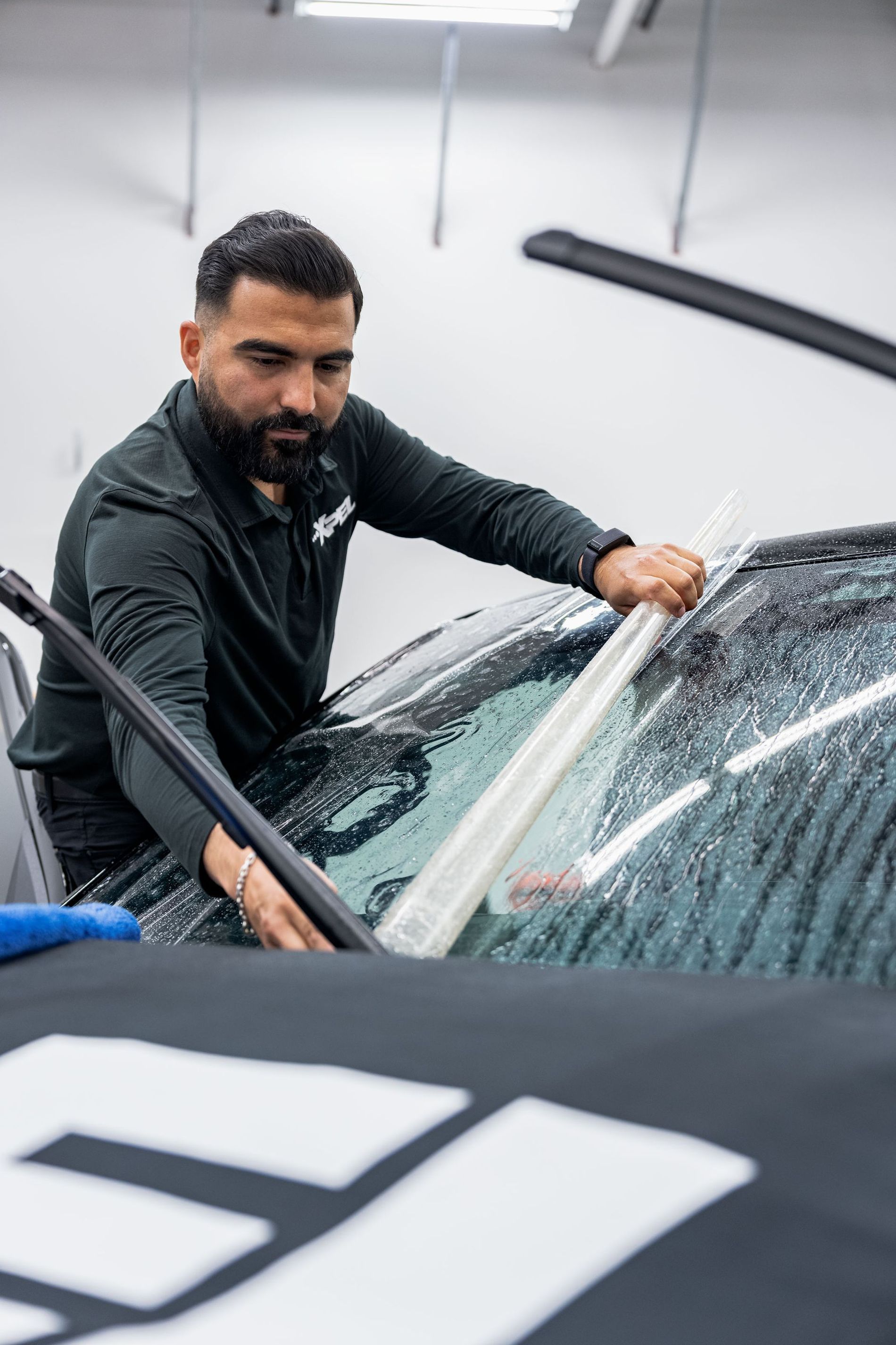 Man applying film to a car's windshield in a shop. He wears a black shirt and focuses on his work.