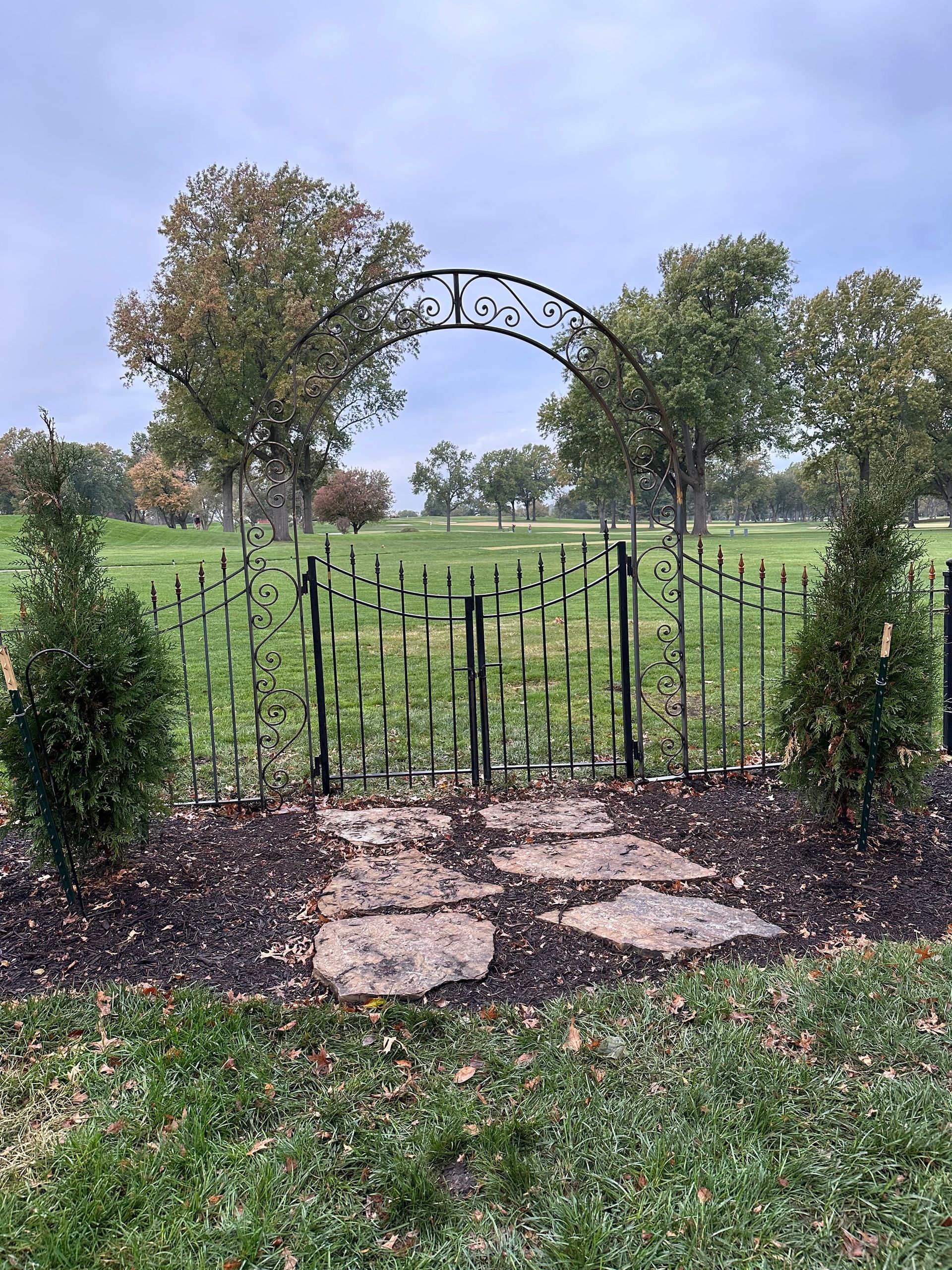Black iron garden gate and archway, stone path, green lawn, fall trees in background.