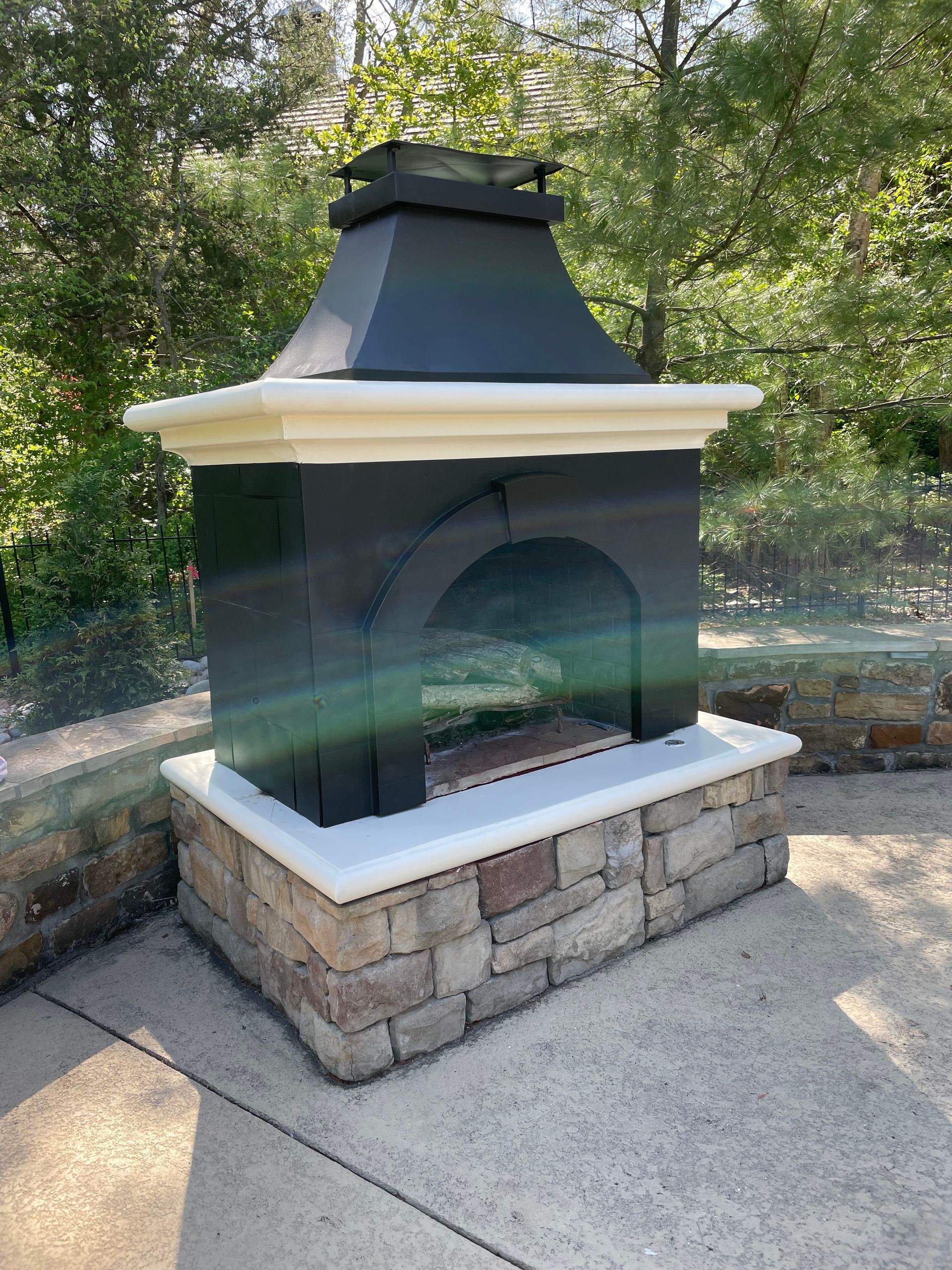 Outdoor fireplace with stone base, black body, and white trim against a backdrop of trees.