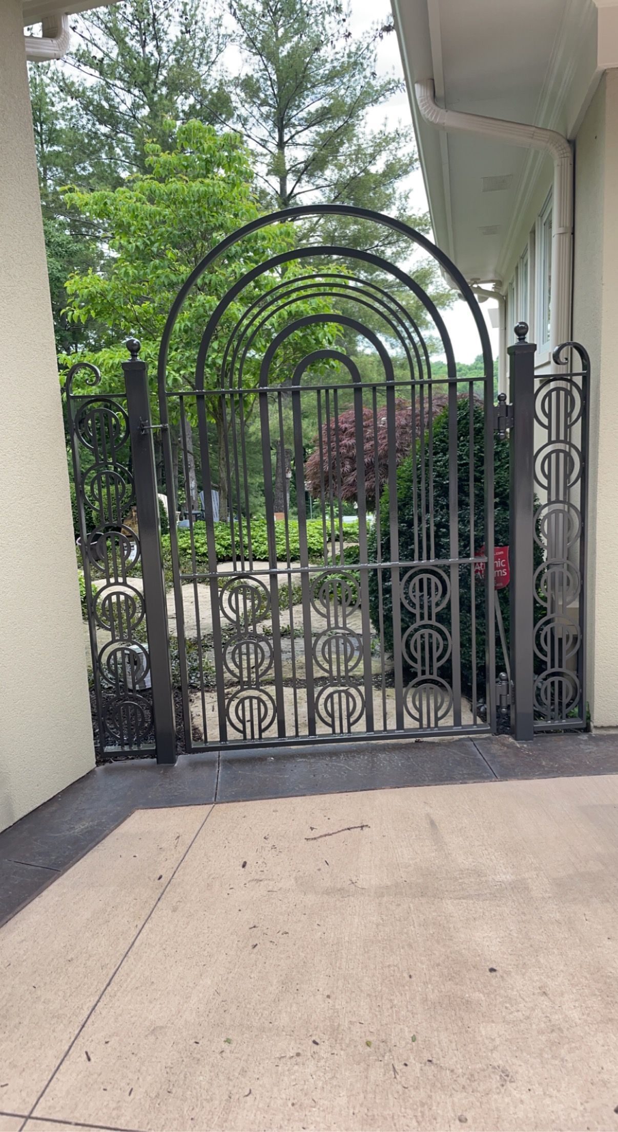 Black iron garden gate with arched top, opening to a path lined with plants and trees.
