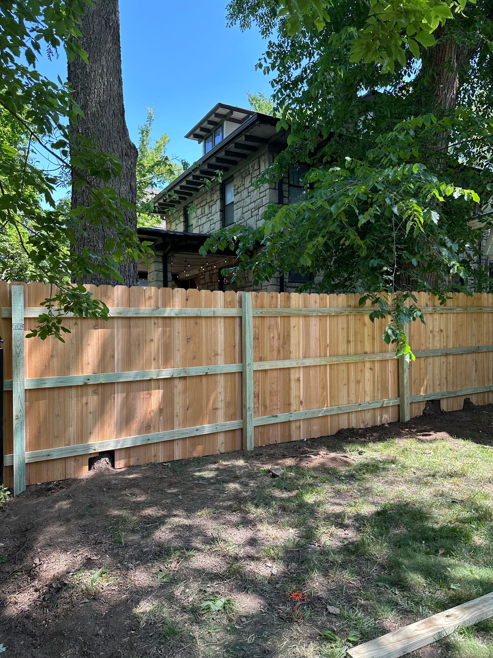 Wooden fence in a yard, partially obscuring a two-story stone house surrounded by trees.