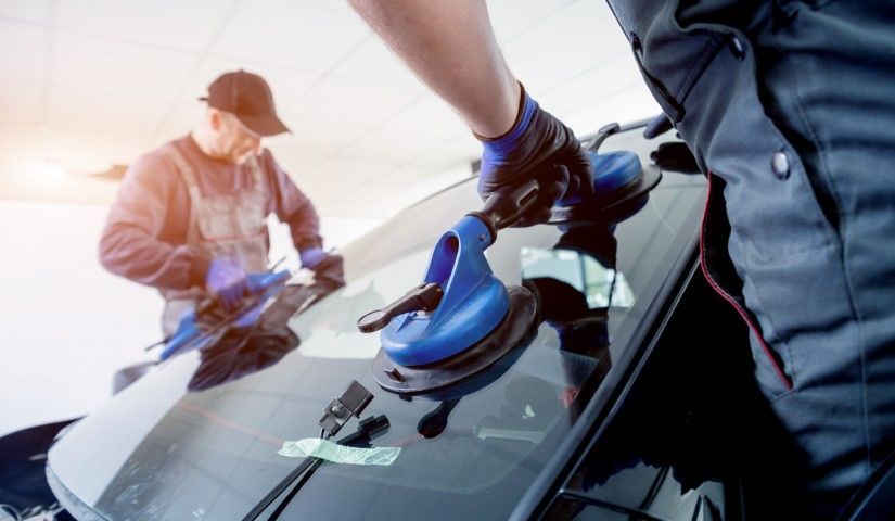 Workers polishing a car windshield, one using a blue suction tool on the hood outdoors