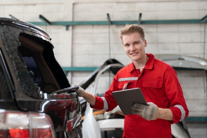 Mechanic in red uniform inspecting a black car in a garage, holding a clipboard.