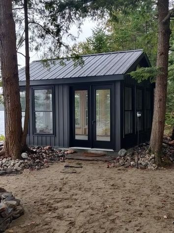 Small black cabin with a metal roof in a wooded area, surrounded by trees and rocks.