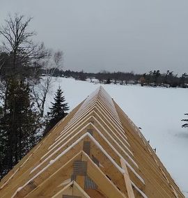 Snow-covered roof trusses of a building under construction overlooking a frozen lake on a gray day
