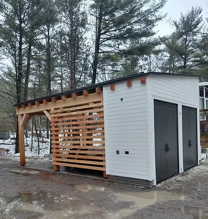 Small white shed with a wood slatted pergola, set on a wet wooded lot in winter.