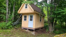 Small wooden tiny house with white door and windows in a wooded clearing