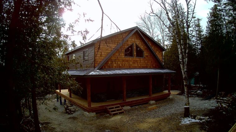 Rustic wooden cabin with a covered porch in a wooded yard, framed by trees and bare branches.