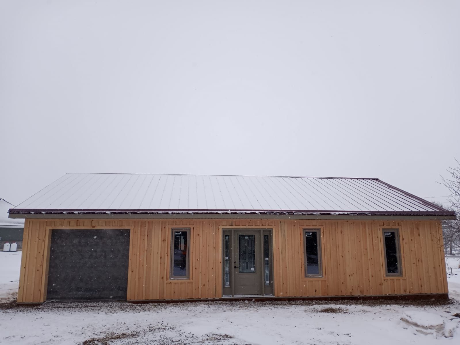Snow-covered wooden house with a garage door and two windows under an overcast sky