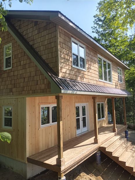 Two-story cedar-clad house with porch, large windows, and sloped roof in a wooded setting