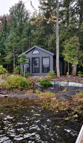 Small gray cabin by a forested lakeshore, with dark water in the foreground.