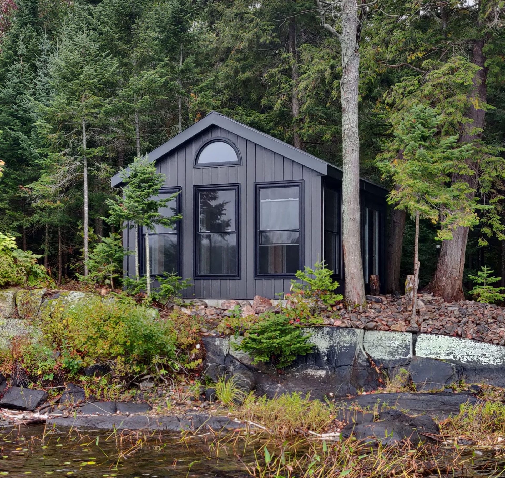 Small gray cabin in a wooded setting beside a rocky shoreline