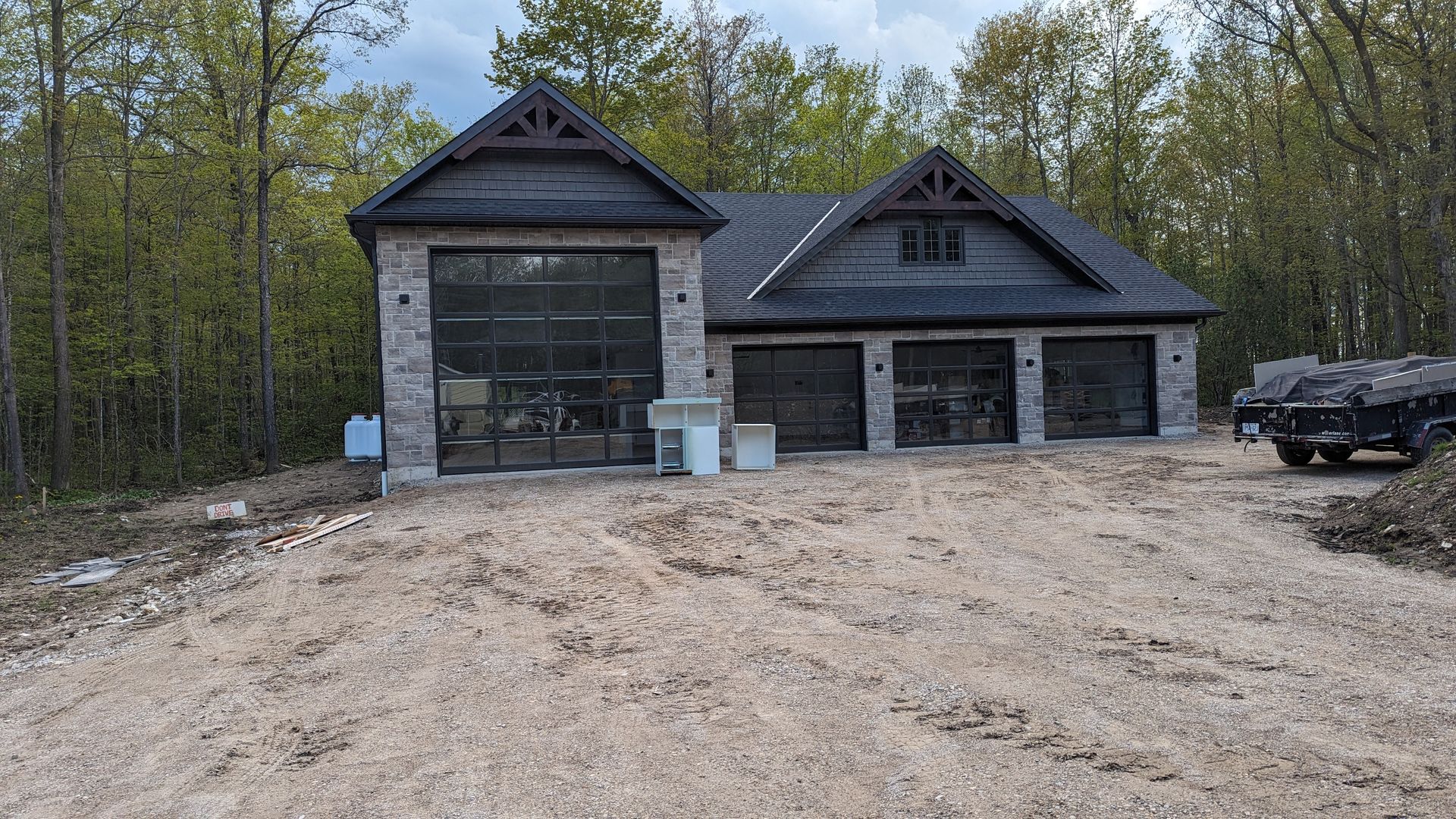 New gray house with attached garage on a dirt lot, backed by trees and a parked truck