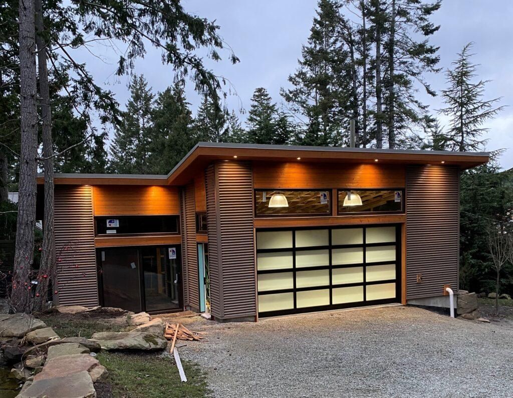 Modern wood cabin garage with lit exterior, glass door, and tall trees in a gravel driveway at dusk