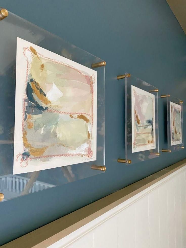 Tres obras de arte abstracto en marcos de acrílico transparente con herrajes dorados cuelgan de una pared azul.