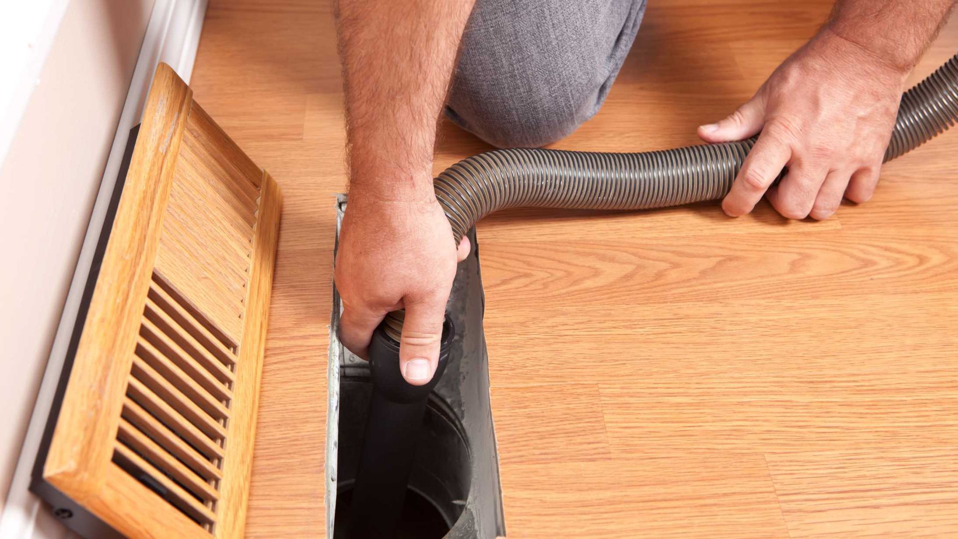Hands using a vacuum hose to clean a wooden floor beside a vent or floor register.