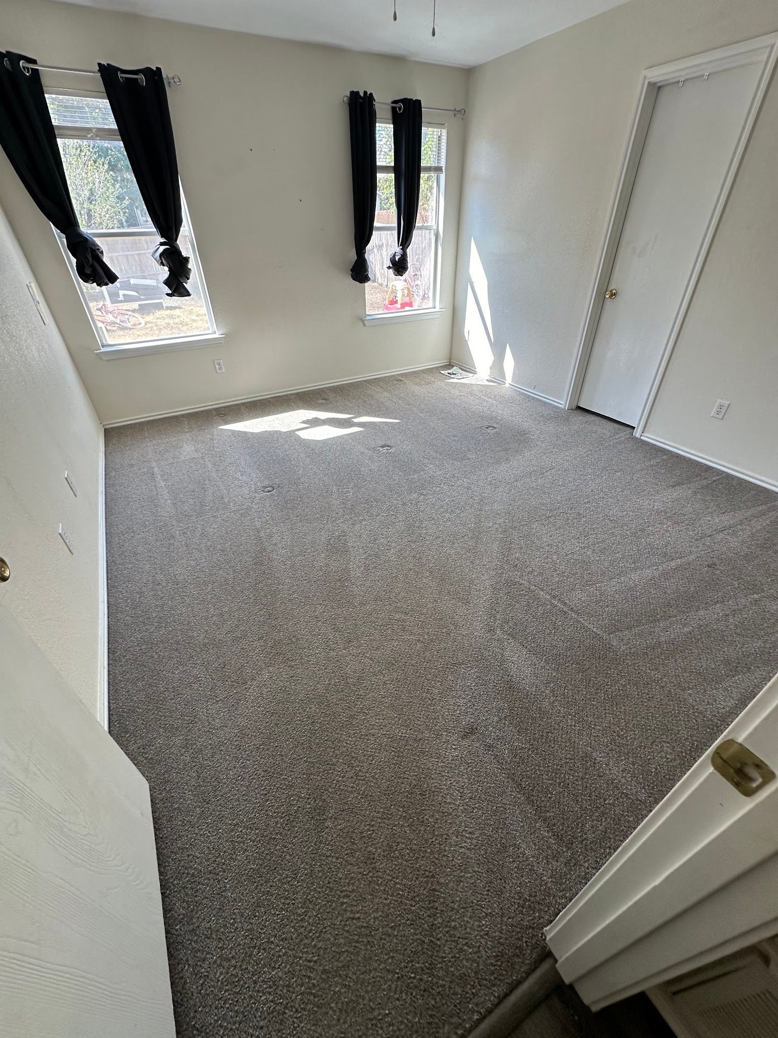 Empty beige bedroom with gray carpet, two black-curtained windows, and white closet doors.