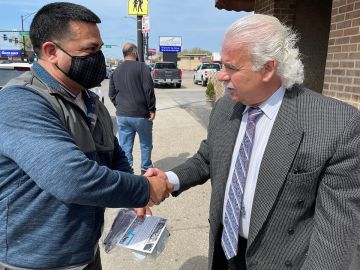 Two men shaking hands on a sidewalk in front of a building. One man is wearing a mask, the other a suit.