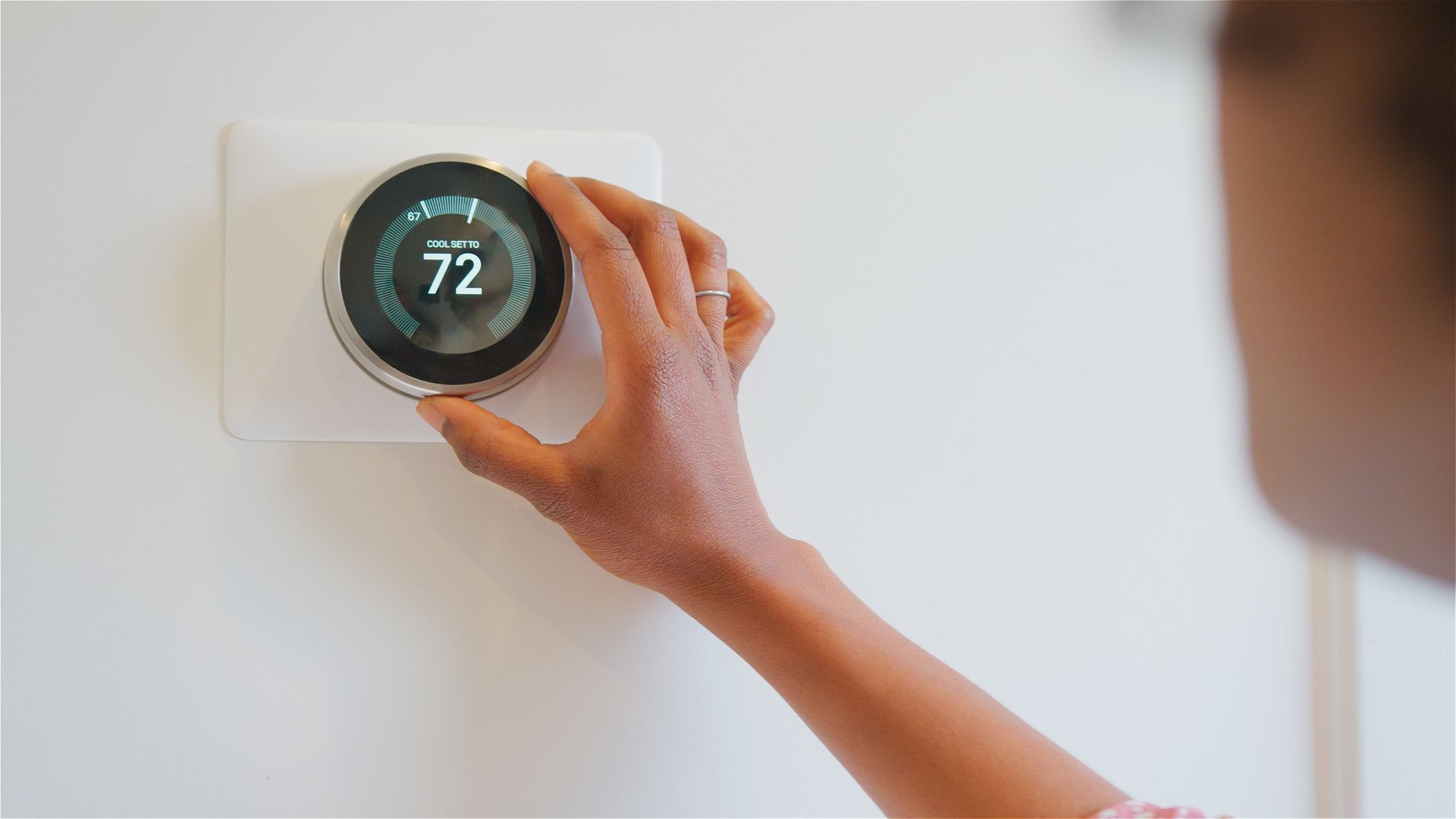 Hand adjusting a round smart thermostat on a white wall, showing 72 degrees.