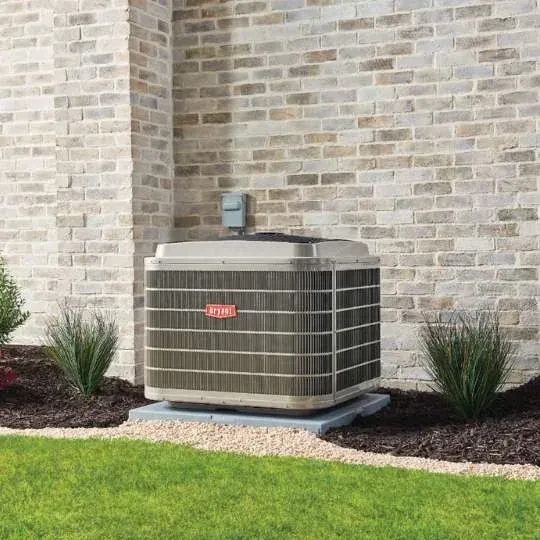 Outdoor air conditioner unit beside a brick house wall, surrounded by mulch and grass.