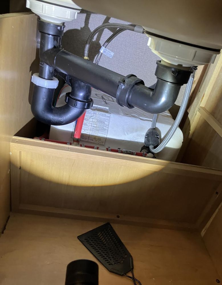 Under-sink cabinet with exposed plumbing pipes and a black dustpan on the wooden floor panel.