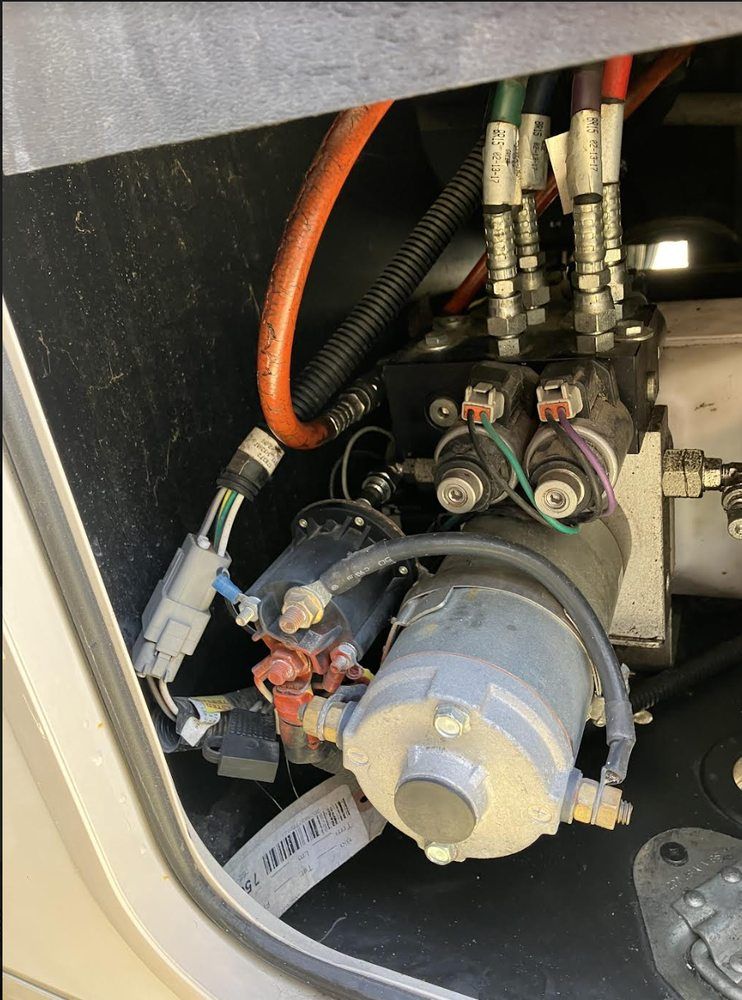 Hydraulic pump assembly with various colored hoses and electrical wiring, mounted in a vehicle compartment.