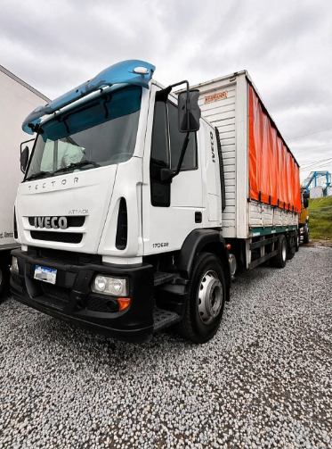 Camión Iveco blanco con remolque cubierto con una lona naranja estacionado en un terreno de grava bajo un cielo nublado.
