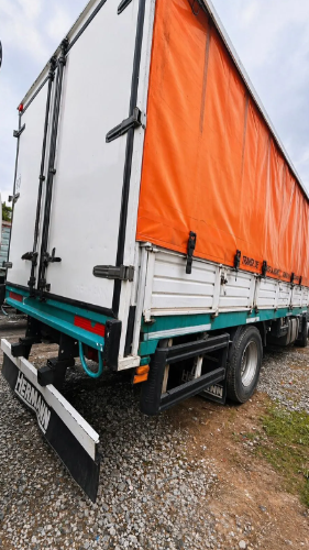 Camión Iveco blanco con remolque cubierto con una lona naranja estacionado en un terreno de grava bajo un cielo nublado.