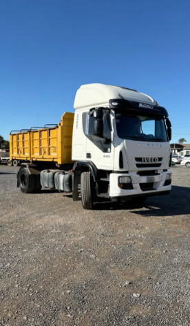 Camión semirremolque blanco con caja basculante amarilla estacionado sobre grava bajo la luz del sol.