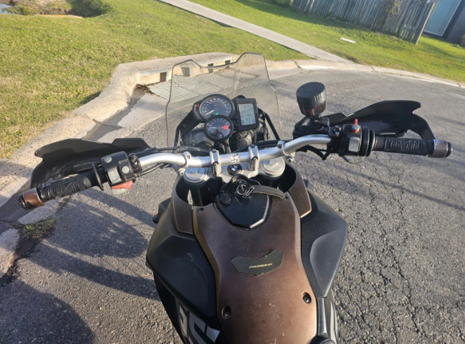 Vista del tablero y el parabrisas de una motocicleta de una carretera curva junto a un parque cubierto de hierba.