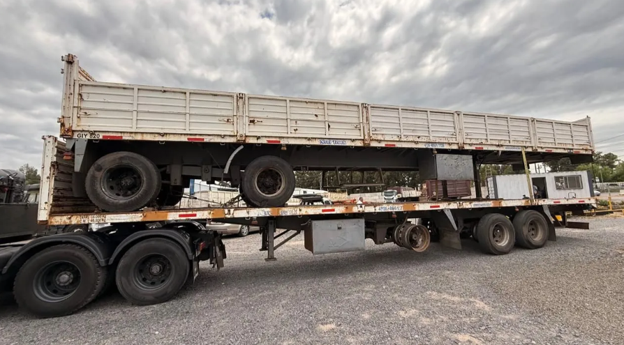 Semirremolque de plataforma que transporta un remolque de carga blanco en un terreno de grava bajo un cielo nublado.