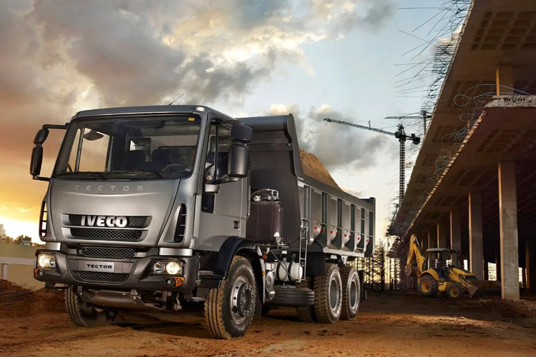 Camión volquete Iveco gris en un camino de tierra en una obra en construcción cerca de un edificio sin terminar al atardecer.