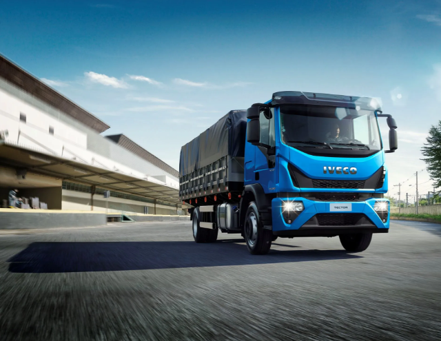 Camión de carga Iveco azul circulando por una carretera junto a un almacén moderno.