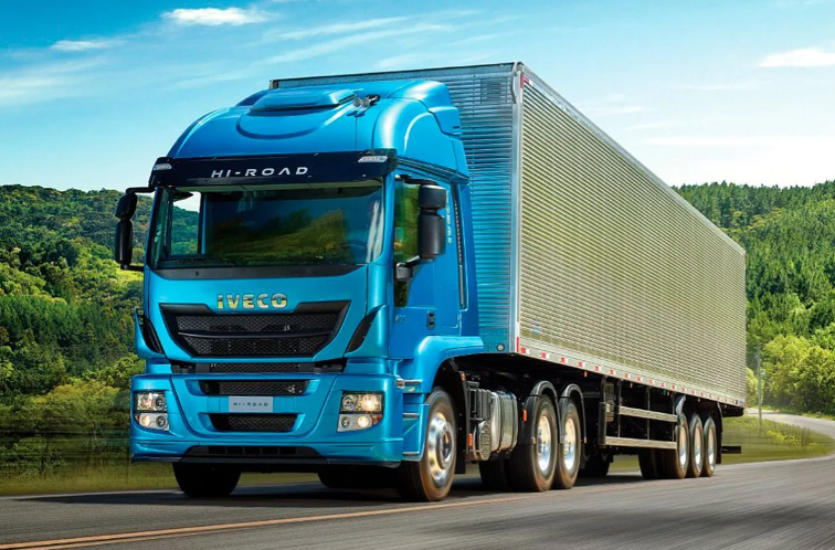 Un camión semirremolque Volvo azul circula por una autopista con un largo remolque plateado bajo un cielo azul.