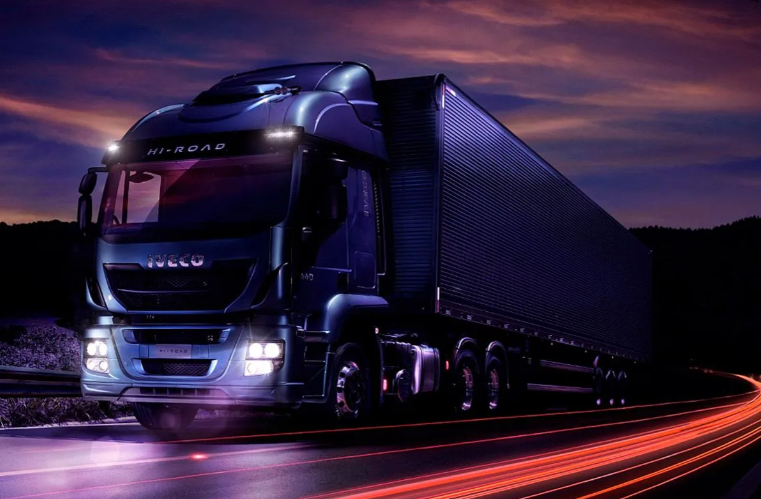 Un camión semirremolque Volvo azul circula por una autopista con un largo remolque plateado bajo un cielo azul.