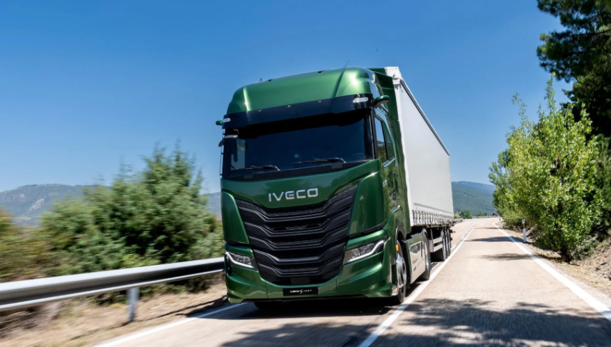 Un camión semirremolque Iveco verde circula por una carretera soleada con montañas y árboles al fondo.
