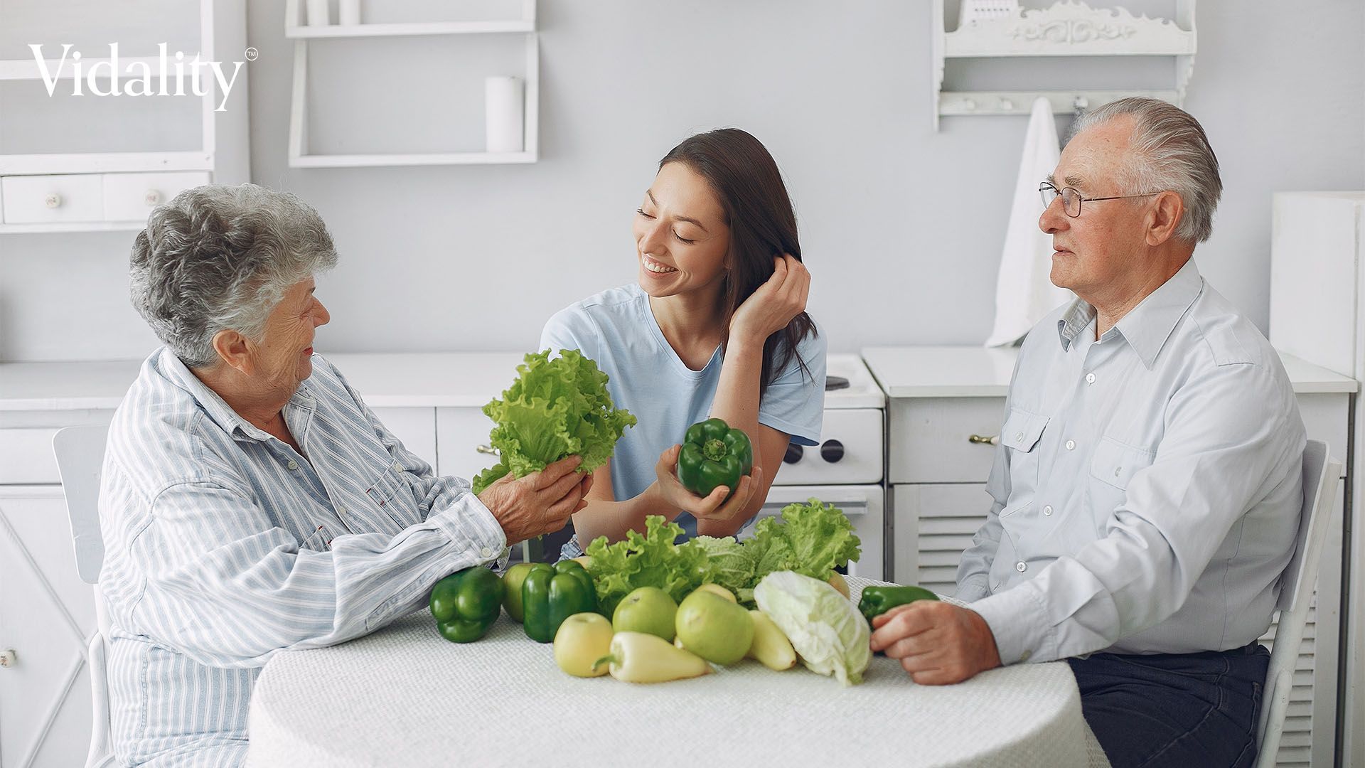 An elderly couple and a caregiver sitting at a table with vegetables, enjoying a healthy meal together.
