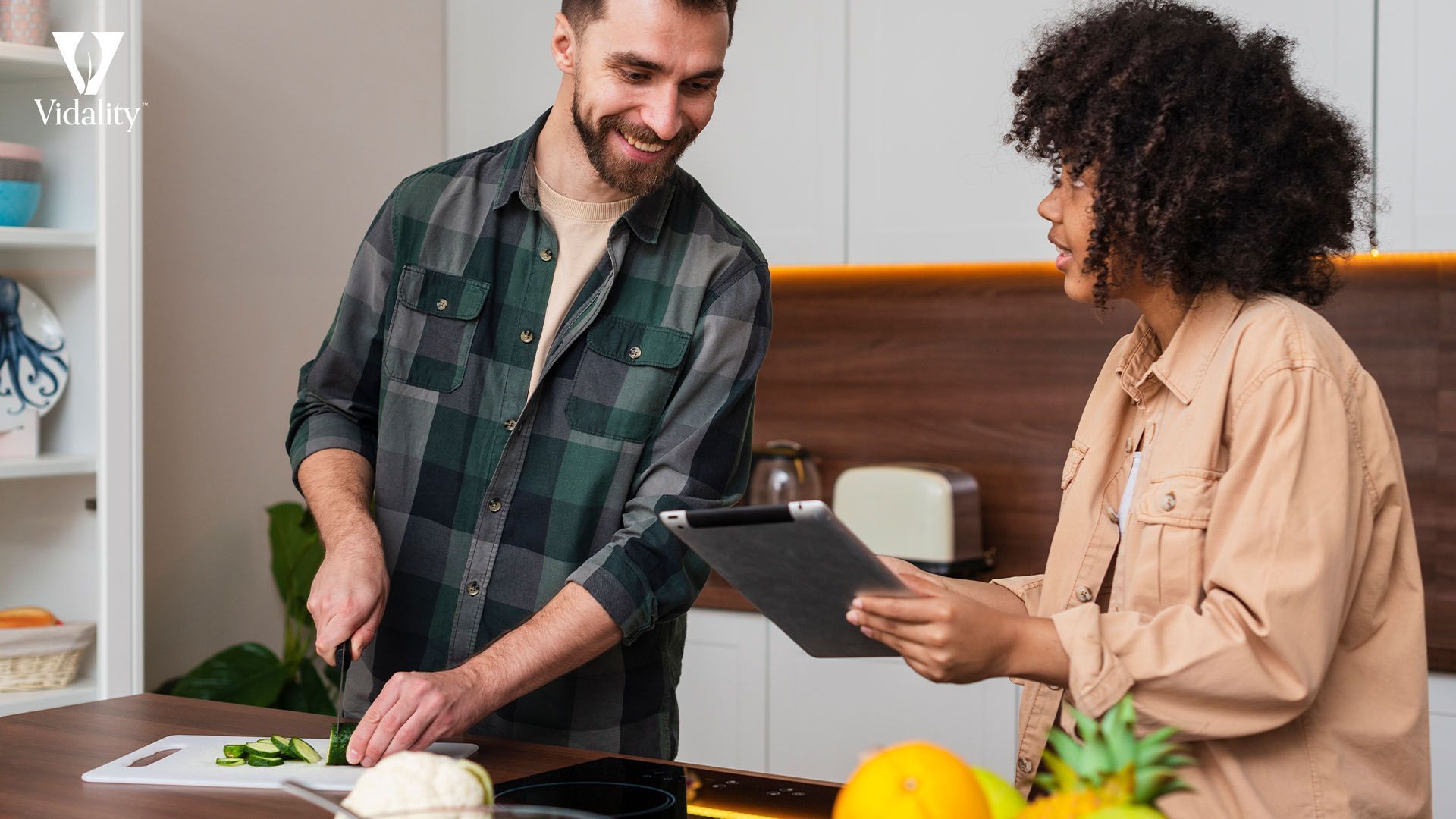 A man and woman stand in the kitchen, looking at a tablet.