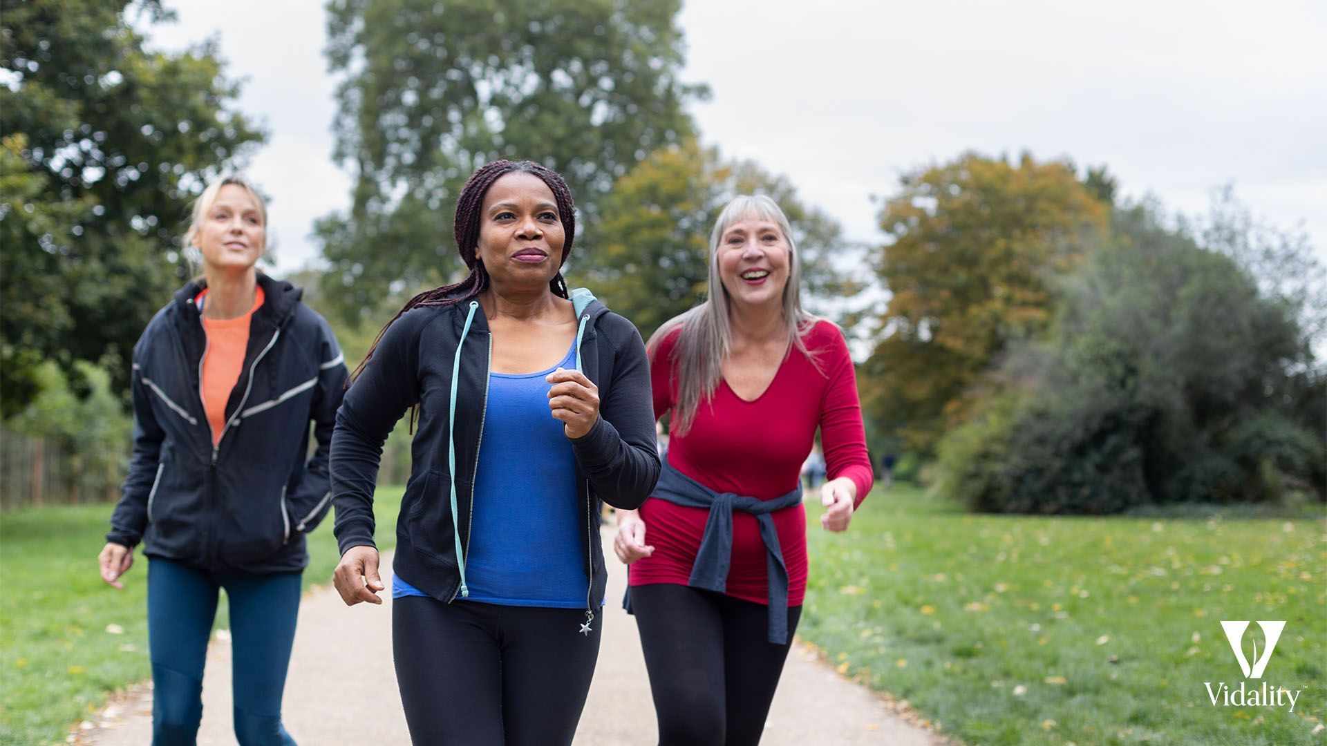 Three women jogging in a park on a sunny day, wearing athletic clothing and headphones, surrounded by green trees and a clear blue sky.